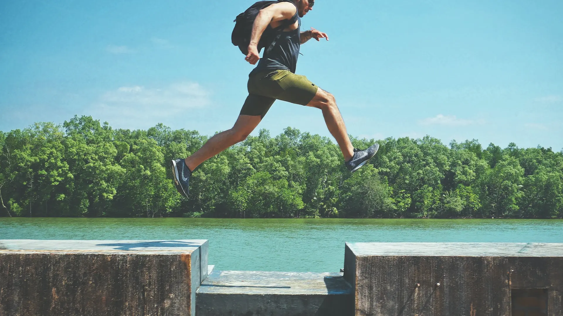 man leaping on concrete surface near body of water and forest at the distance during day
